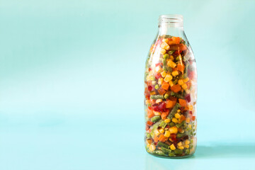 Frozen vegetables in a transparent glass bottle on a light blue background. The concept of using quick-frozen products