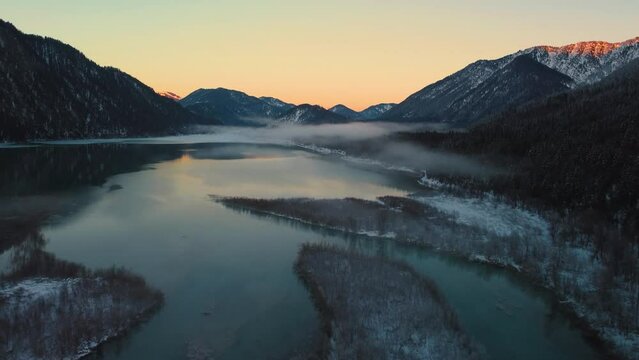 Scenic mountain river valley with fresh water in the Bavarian Austrian Alps at Sylvensteinspeicher by sunshine sunset, winter snow riverbed along trees and forest and mountains. Drone aerial flight.