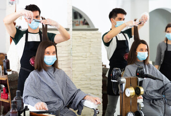 Fototapeta na wymiar Positive male hairdresser in protective mask and gloves making hair styling for young woman, working day in beauty salon during pandemic situation
