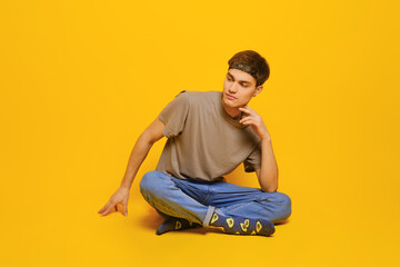 Cheerful young man in casual clothes sitting on the floor in bright yellow studio