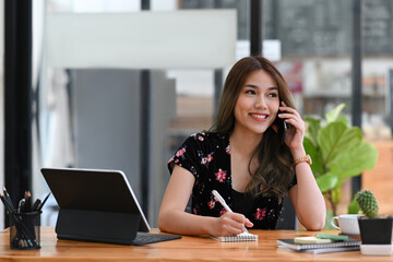 Smiling young asian woman talking on mobile phone and making notes on notebook.