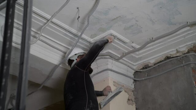 Fat bearded adult man in protective helmet and black tight fitting clothing remove construction whitewash vintage high ceiling. Overhaul repair with your own hands concept. 4k slow motion footage 