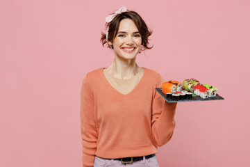 Young smiling cacuasian happy fun cool woman 20s wearing casual clothes holding in hand makizushi sushi roll served on black plate traditional japanese food isolated on plain pastel pink background