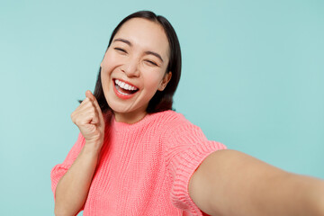 Close up young happy woman of Asian ethnicity 20s in pink sweater do selfie shot pov on mobile phone do winner gesture isolated on pastel plain light blue background studio. People lifestyle concept.