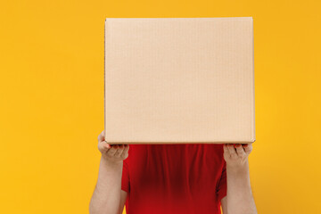 Professional delivery guy employee man wearing red cap T-shirt uniform workwear work as dealer courier hold cover face with cardboard box isolated on plain yellow background studio. Service concept.