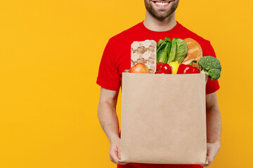 Cropped delivery guy employee man 20s wear red cap T-shirt uniform workwear work as dealer courier hold craft brown paper bag with grocery food vegetables isolated on plain yellow background studio