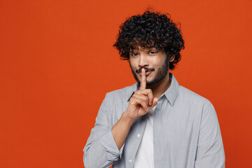 Fototapeta na wymiar Secret calm fascinating young bearded Indian man 20s years old wears blue shirt say hush be quiet with finger on lips shhh gesture looking camera isolated on plain orange background studio portrait.
