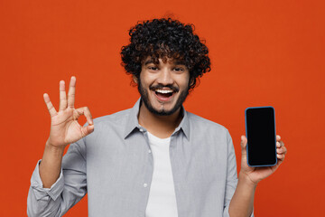 Obraz na płótnie Canvas Young bearded Indian man 20s years old wears blue shirt hold in hand use mobile cell phone with blank screen workspace area showing okay ok gesture isolated on plain orange background studio portrait.