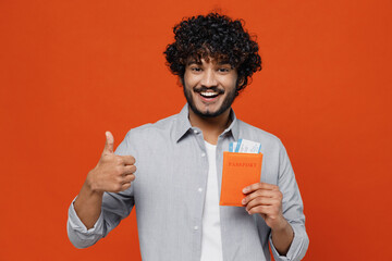 Confident excited fun exultant young bearded Indian man 20s years old wears blue shirt hold passport boarding tickets showing thumb up like gesture isolated on plain orange background studio portrait.