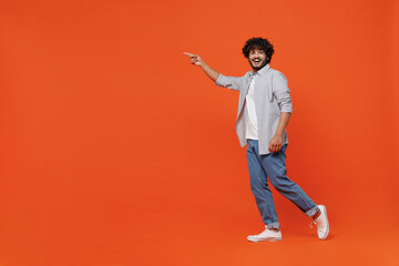 Full size body length side profile view happy young bearded Indian man 20s years old wears blue shirt pointing on workspace area copy space mock up isolated on plain orange background studio portrait.