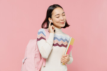 Satisfied happy teen student girl of Asian ethnicity wear sweater backpack hold books listen music in earphones isolated on pastel plain light pink background Education in university college concept