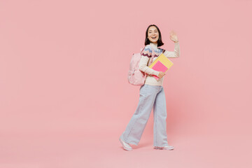 Full body side view teen fun student girl of Asian ethnicity wearing sweater hold backpack book walk going waving hand isolated on pastel plain pink background Education in university college concept