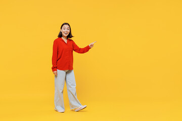 Fototapeta premium Full size body length happy young woman of Asian ethnicity 20s in casual clothes look camera go move pointing on workspace area copy space mock up isolated on plain yellow background studio portrait.