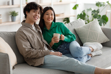 Obraz premium Happy weekend together. Affectionate young Asian couple drinking coffee and cuddling on cozy couch at home