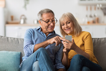 Older Spouses Using Mobile Phone Sitting On Sofa At Home