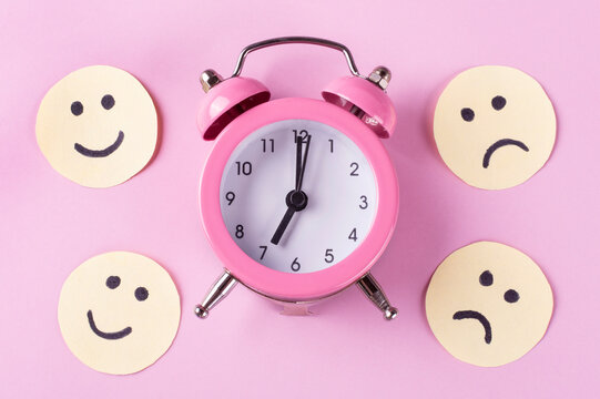 Mechanical classic alarm clock (hands at 7 o'clock in the morning), funny and sad emoticons on a pink background. The concept of early awakening (wake up). Good and bad moods in the morning.