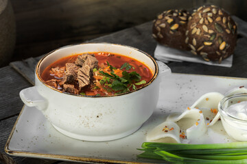 red borscht with beef in a white plate stands on a wooden box, served with bacon, green onions, mustard in a glass bowl, sour cream in a glass bowl, rye buns with sunflower seeds, rustic style l