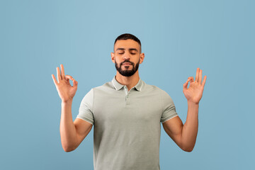 Stress relief concept. Middle eastern man meditating with closed eyes, keeping calm on blue studio...