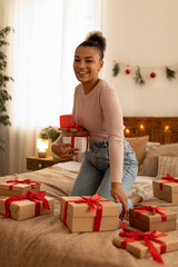 Excited african american woman holding presents in hand and smiling while sitting on bed surrounded by Christmas gifts