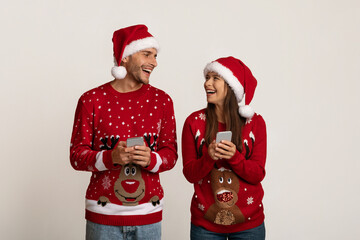 Cheerful Couple In Santa Hats Holding Smartphones And Looking At Each Other