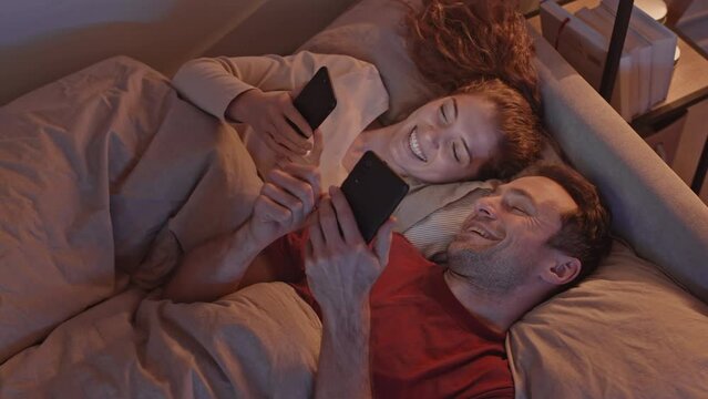 Top view slowmo shot of young Caucasian couple using smartphones and chatting while lying together in bed at night