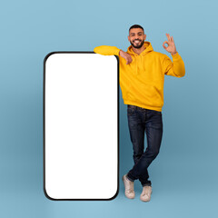 Excited arab man showing okay gesture, standing next to huge smartphone over blue background. Advertisement mockup