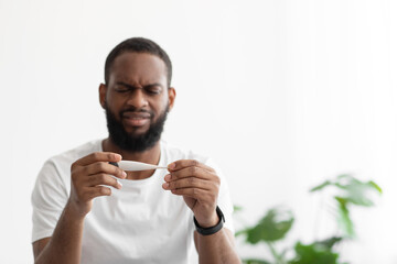 Unhappy sad young black bearded man hold thermometer with high temperature in bedroom