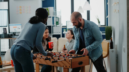 Diverse group of workmates enjoying game with foosball table to do fun activity after work. Coworkers playing football and drinking beer from bottles, celebrating party with entertainment