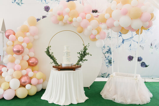 A large three-tiered white cake for the baptism of a child with green twigs on the background of a beautiful photo zone with balloons and a candy bar
