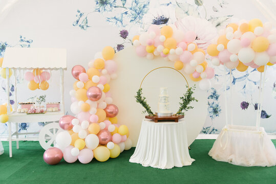 A large three-tiered white cake for the baptism of a child with green twigs on the background of a beautiful photo zone with balloons and a candy bar