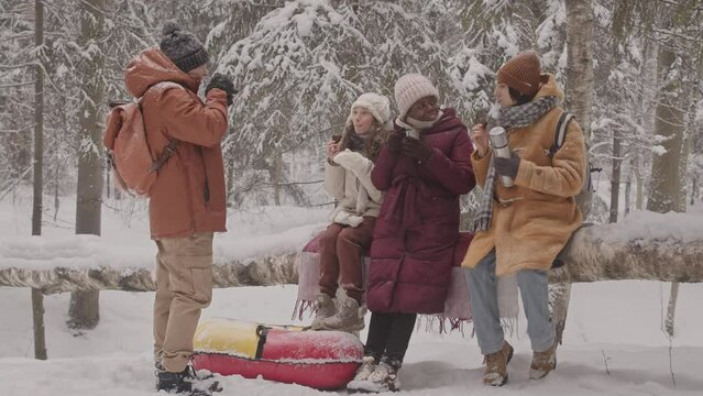 Slowmo shot of four young multiethnic friends spending day in beautiful winter forest, drinking hot tea from thermos and taking pictures of themselves on smartphone