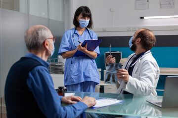 Physician talking to nurse and holding x ray scan at checkup visit with retired man in office. Doctor explaining radiography diagnosis to patient while he asks medical assistant to help.