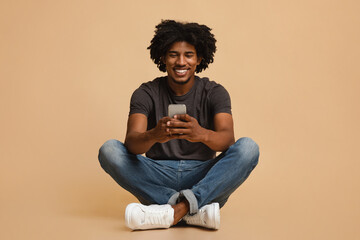 Happy millennial african american guy using smartphone, sitting over beige studio background