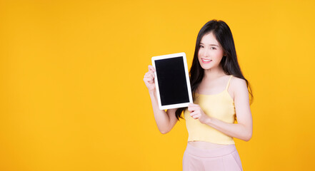 Asian employee woman showing screen tablet with blank monitor over isolated. Cheerful asia student girl holding digital tablet with smile on face on yellow background. Technology digital concept.