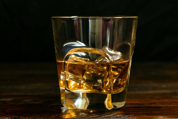 A close-up of a half-poured glass of whiskey with ice cubes on the dark oak boards.