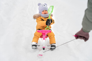 Toddler 12-17 months old with a green toy shovel in his hands rides a children's snowcat which is pulled by one of the parents by a rope. Active lifestyle in winter