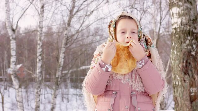 Cute girl in a traditional Russian headscarf with bagels eats pancake on winter background. Closeup portrait of a child in folk clothes. Maslenitsa festival. Traditional Russian food for Shrovetide.
