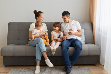 Happy family sitting on sofa in living room wearing casual clothing, spending weekend together, using cell phones, expressing positive emotions, being glad to spend time together.
