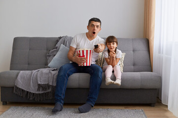 Portrait of young brunette man watching horror movie at home with little daughter while sitting...