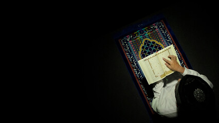 a muslim boy reading quran in a dark room on a prayer matt
