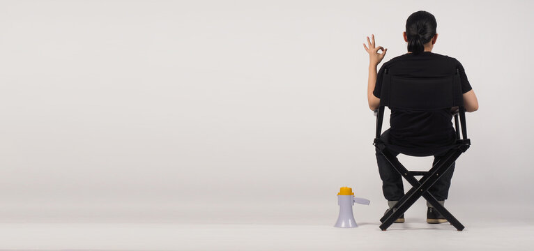 Back of Asian man sitting on black director chair. He doing an A-OK hand sign. Megaphone is put on the floor with a white background.