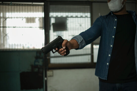 A man in a blue jeans shirt with a mask and a silver cap is standing in the room with a pistol. Point and aim the gun at the target. concept of assassination, murder, criminal