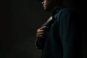 A man in a blue denim shirt is standing in a dark room holding a pistol to his chest. concept of assassination, murder, criminal
