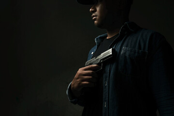 A man in a blue denim shirt is standing in a dark room holding a pistol to his chest. concept of...