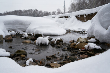 Flowing river with shores covered in snow in a park. Riverbed with rocks in it. Forest in the background.