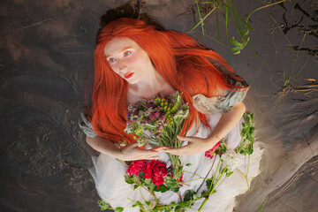 Redhaired girl in a romantic dress lies with open eyes in the water. Young beauty woman with long...
