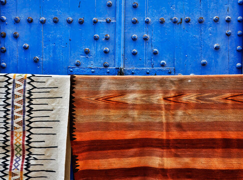 Dos  coloridas alfombras marroqu&iacute;es colgadas ante una cl&aacute;sica puerta azul &aacute;rabe con remaches despintados.