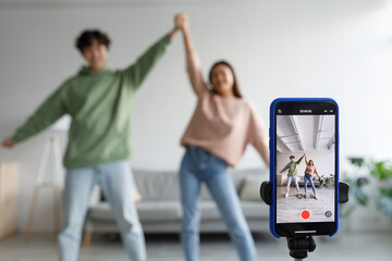 Couple of Asian influencers shooting dance video for social network on cellphone, having fun at home, selective focus