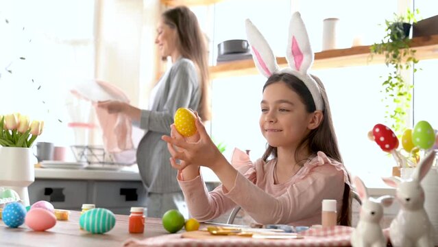 Beautiful cute girl with bunny ears having fun draw, paint, decorate easter eggs. Working on kitchen desk with brushes and paint cartilage, smiling joyfully having fun.  Celebrating spring holiday