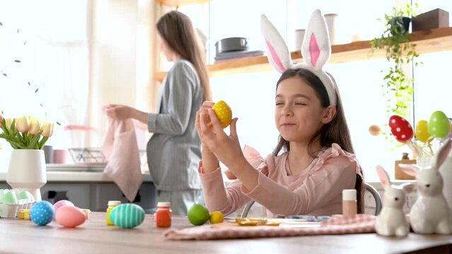 Beautiful cute girl with bunny ears having fun draw, paint, decorate easter eggs. Working on kitchen desk with brushes and paint cartilage, smiling joyfully having fun.  Celebrating spring holiday
